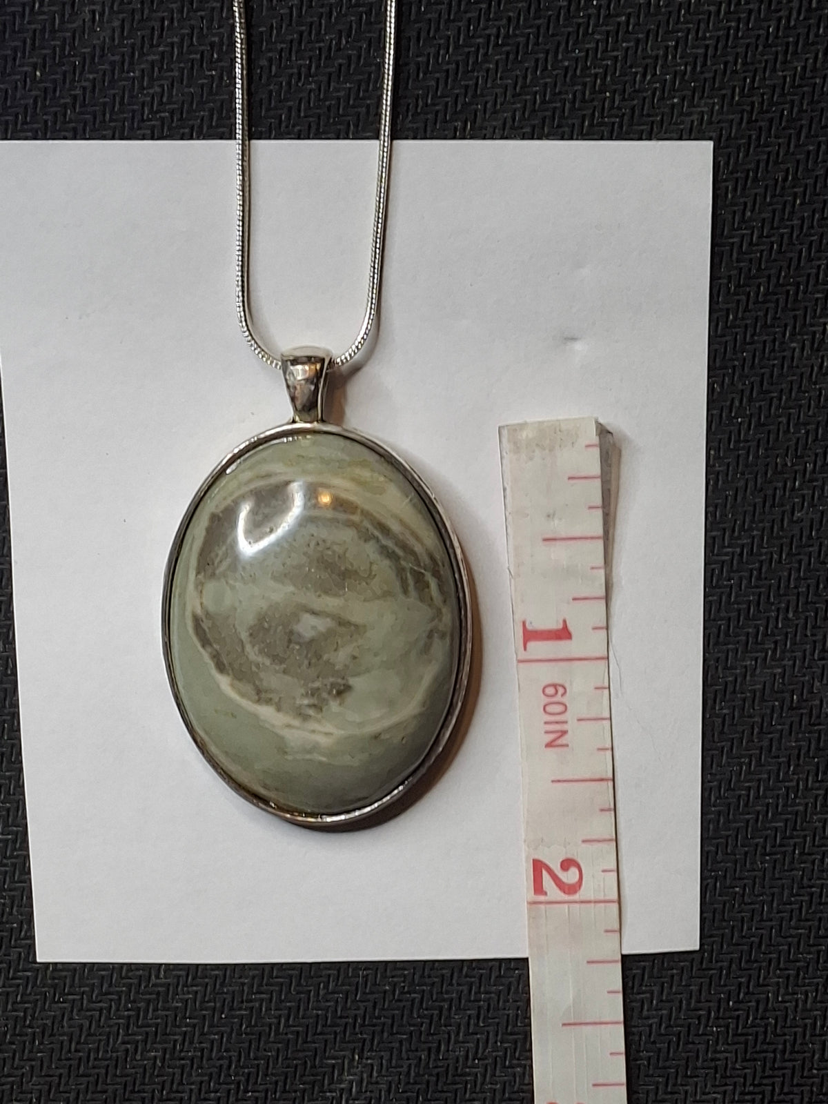 Oval Banded Micaceous Quartzite pendant with silver tone bezel and snake chain, next to a measuring tape on a white paper.