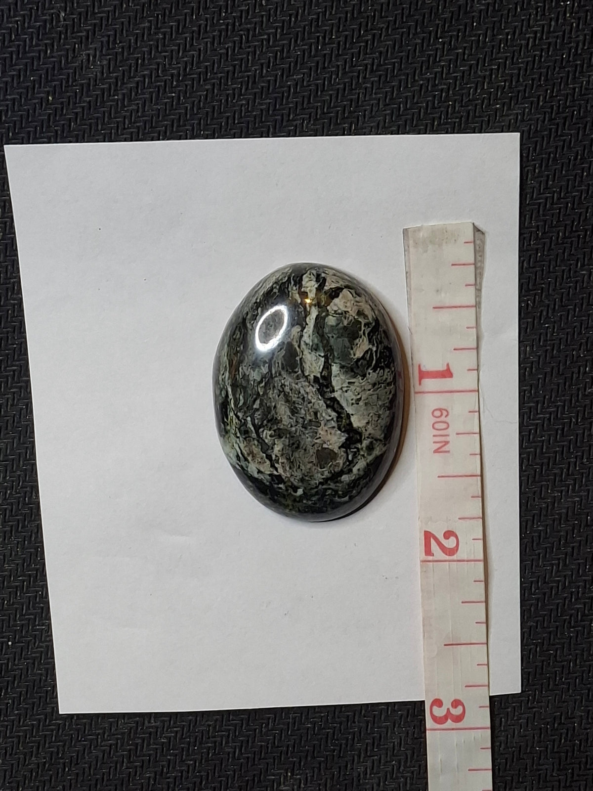 Top-down view of a polished black and grey veined jasper cabochon on white paper with a measuring tape.