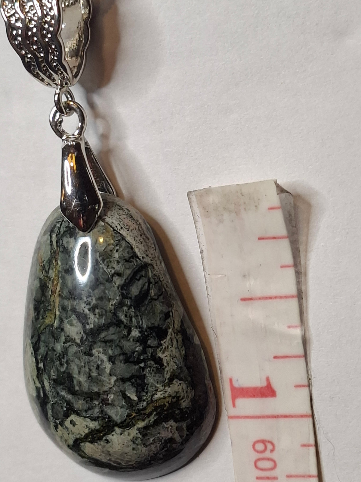 Detailed view of a handcrafted Breccia Aventurine pendant, showing its grey and deep green patterns, with a silver-plated bail and measuring tape.