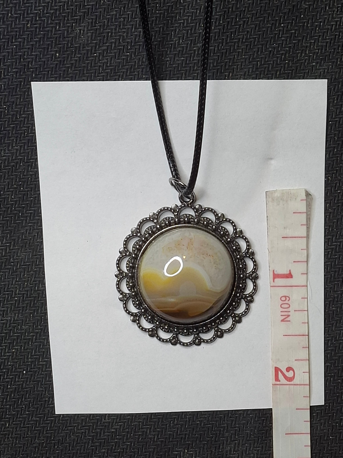 Handcrafted Botswana Agate pendant with a round, domed stone displaying caramel and white bands, set in an ornate dark metal bezel, hanging from a black cord, next to a measuring tape.