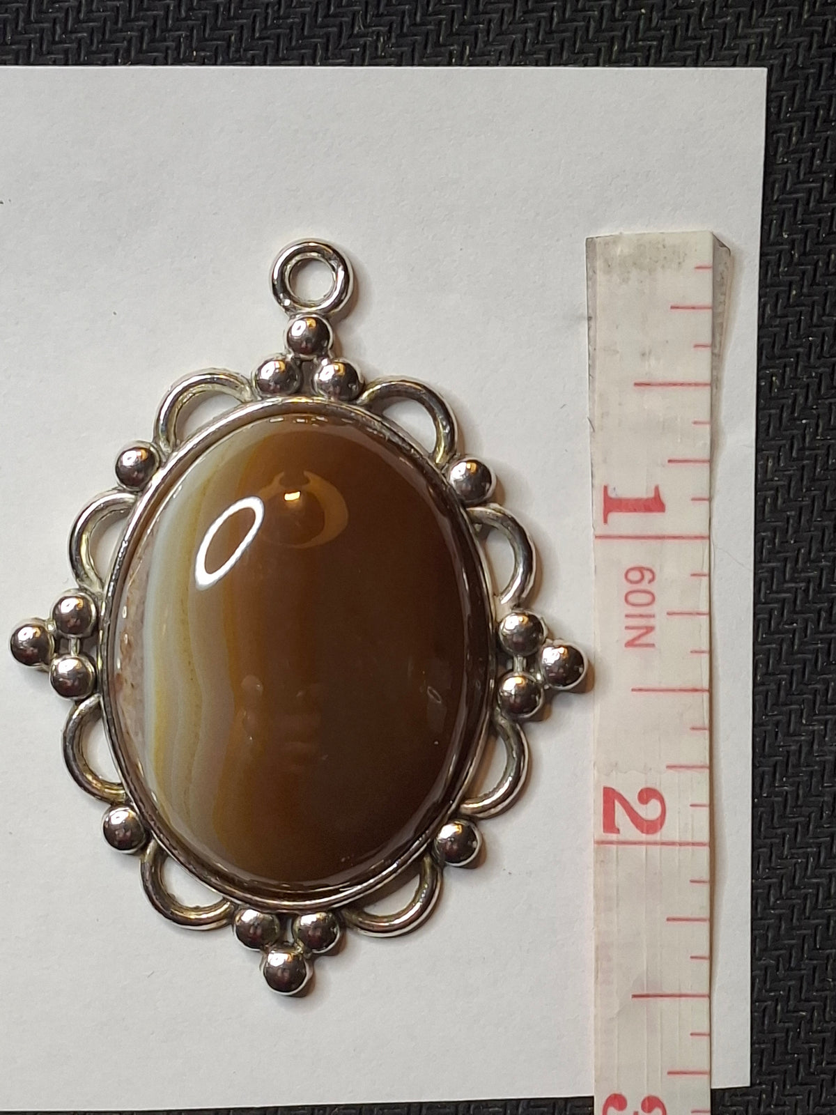Close-up of the oval Botswana Agate pendant with caramel and white bands in an ornate silver-toned setting, with a measuring tape on the right.