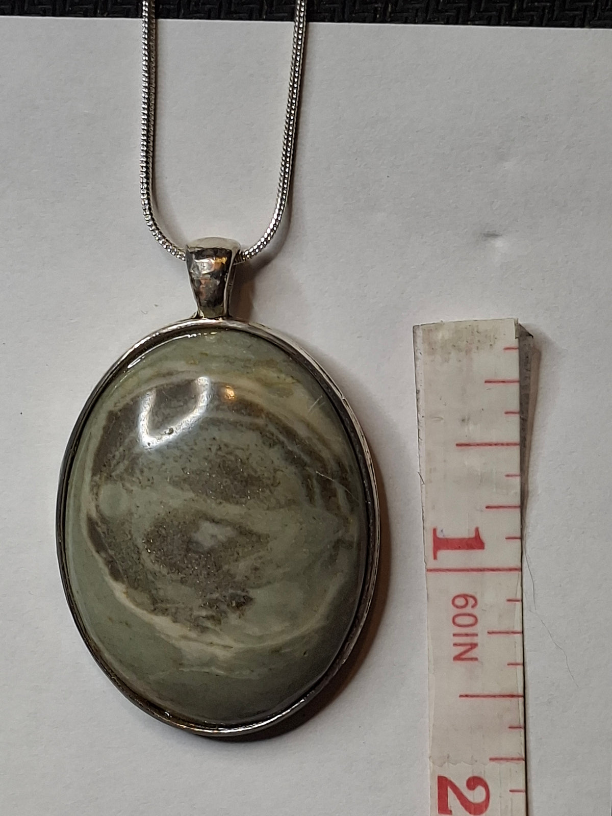 Oval Banded Micaceous Quartzite pendant with silver tone bezel and snake chain, next to a measuring tape, showing swirled grey and cream patterns.