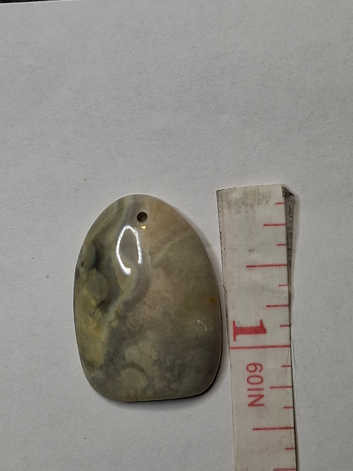 Close-up overhead view of a polished, oval Yellowstone River Jasper pendant stone with a drilled hole, next to a white measuring tape showing its length.
