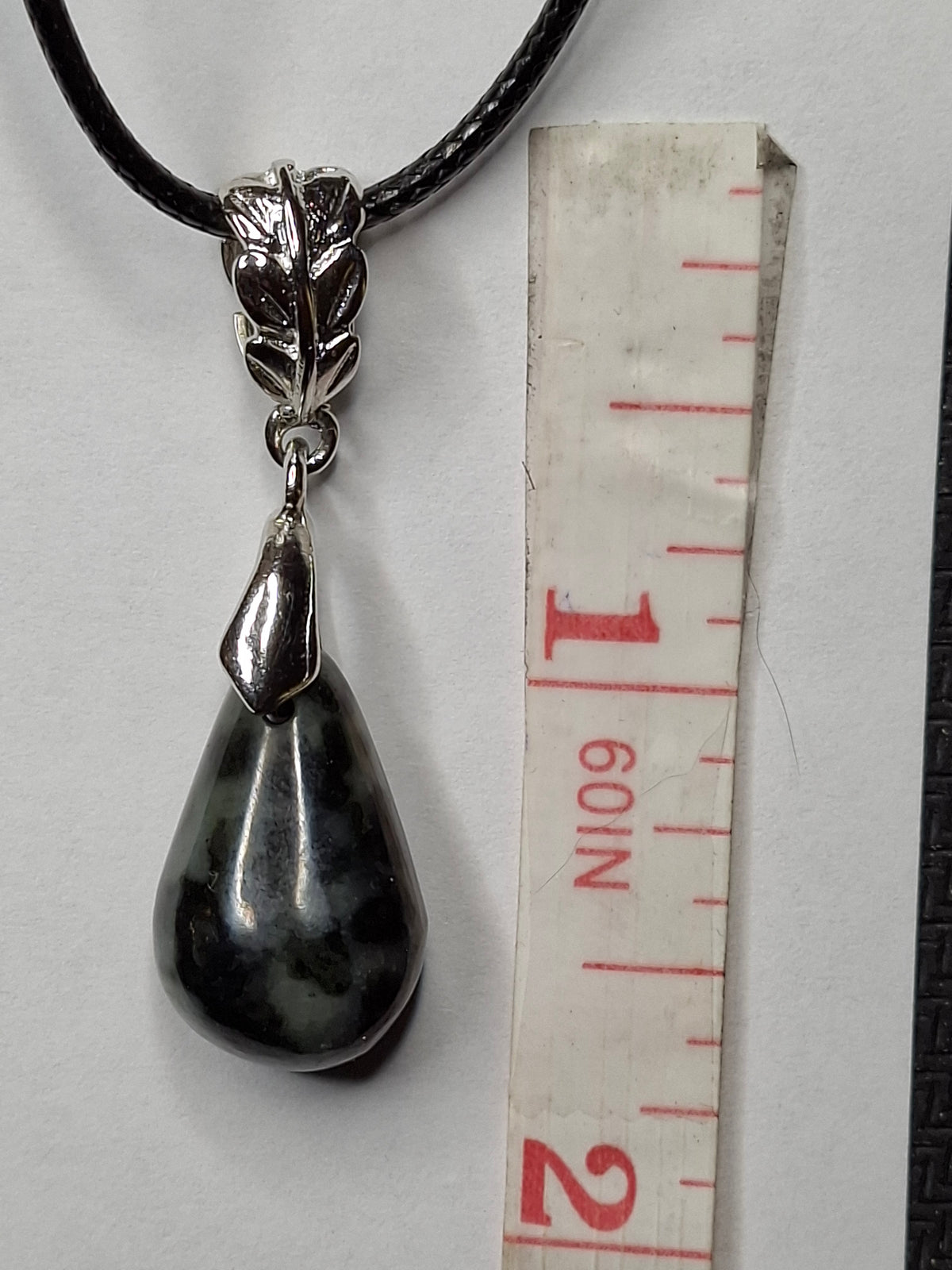 Close-up of a dark green jasper pendant with mottled black spots, featuring a silver-plated leaf-design bail and black cord, next to a white measuring tape showing inches.