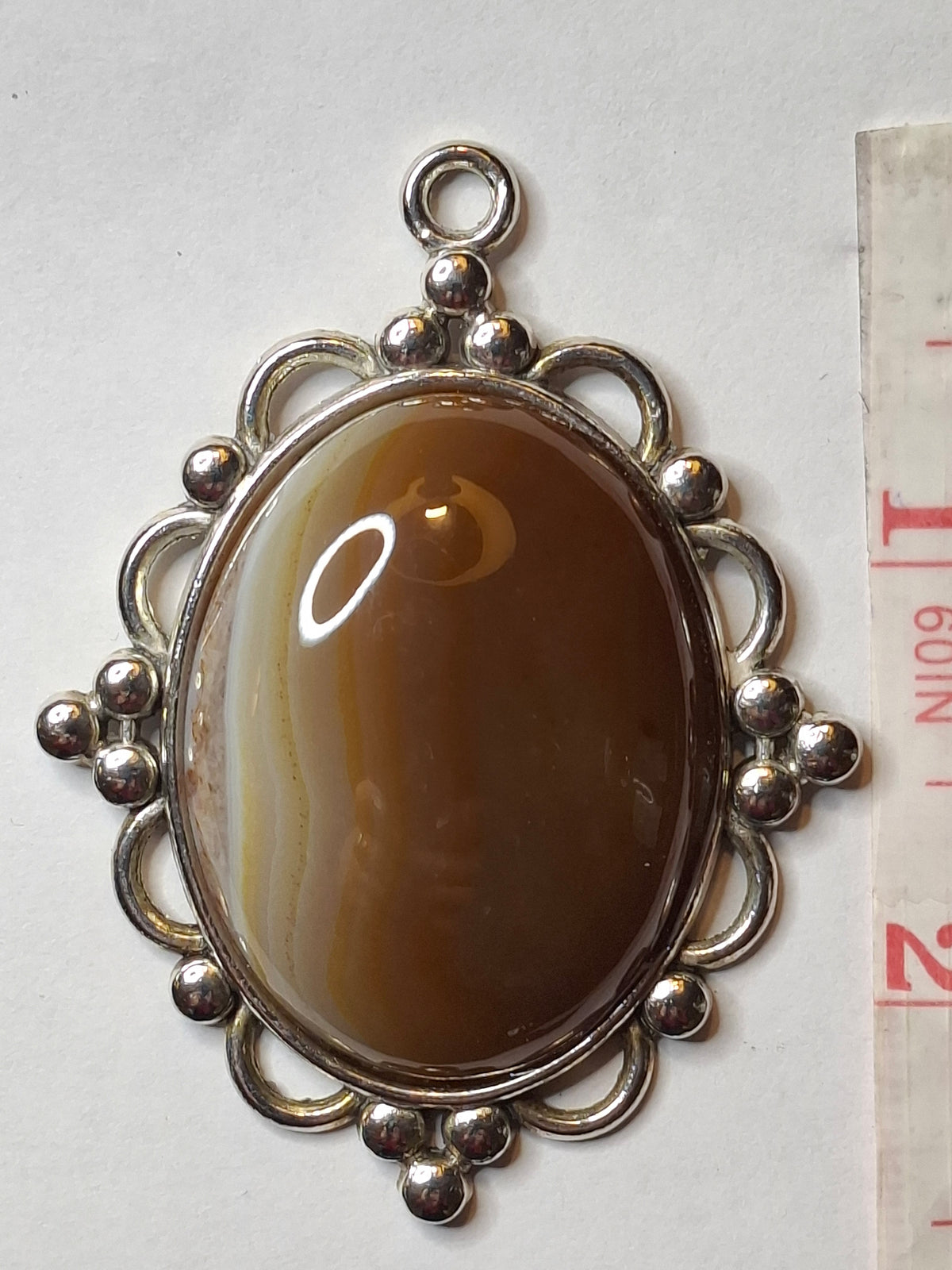 Top-down view of the oval Botswana Agate pendant with caramel and white bands in an ornate silver-toned setting, next to a measuring tape.