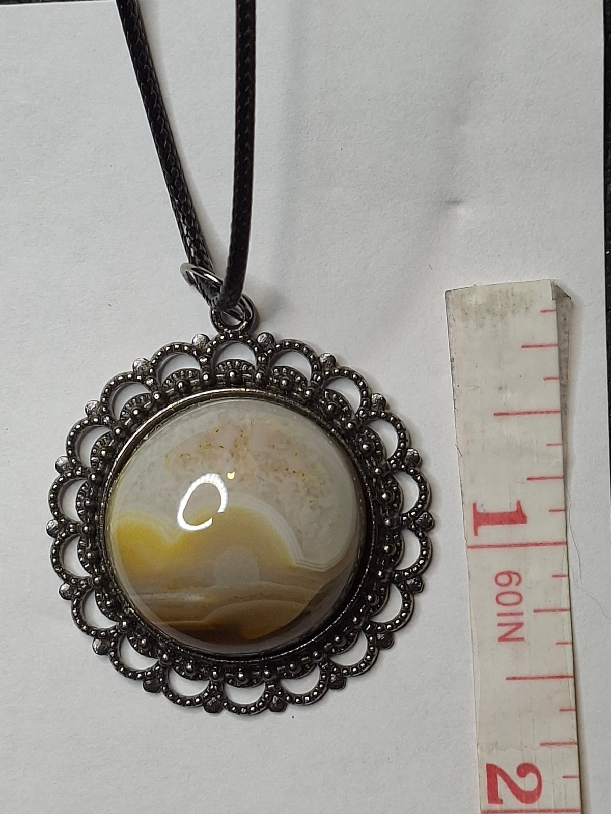 Overhead view of a handcrafted Botswana Agate pendant with a round, domed stone showing caramel and white bands, set in an ornate dark metal bezel, beside a measuring tape on white.