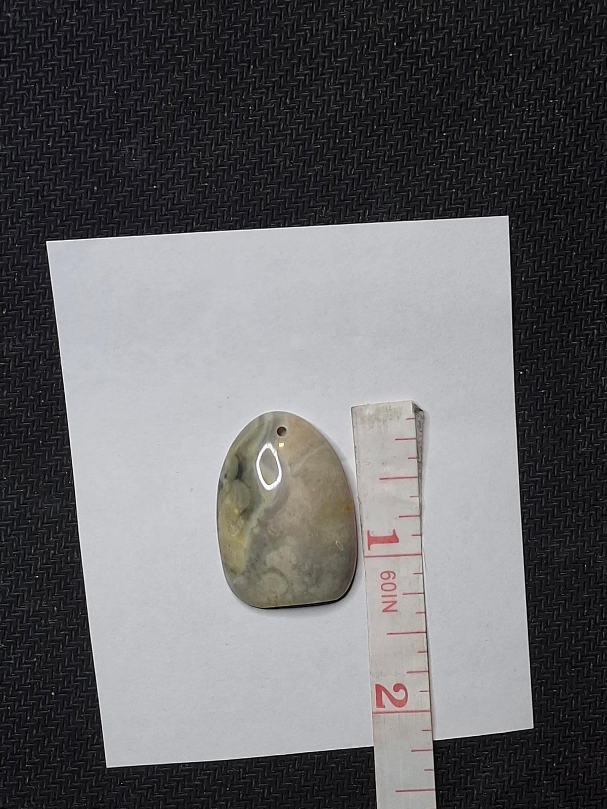 Overhead view of a polished, oval Yellowstone River Jasper pendant stone with a drilled hole, next to a white measuring tape showing its length.