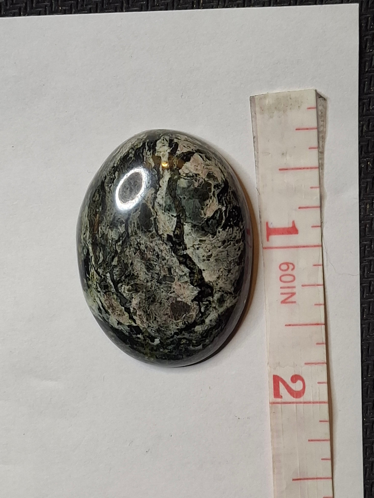Oval black and grey veined jasper cabochon on white paper with a measuring tape on the right.
